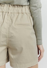 Beige bomullsshorts med elastisk midjebånd, med doble baklommer og en jevn tekstur. Kantede kanter ved bena.