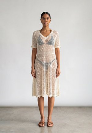 Donna che indossa un vestito di pizzo beige trasparente sopra un bikini nero, in piedi a piedi nudi su un pavimento grigio chiaro uniforme con uno sfondo di parete neutro.