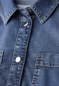Col de chemise en denim bleu avec boutons-pression métalliques et coutures visibles, présentant une poche poitrine sur le côté droit.