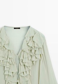 Jasnozielona bluzka z dekoltem w kształcie litery V i wieloma falbaniastymi akcentami na przodzie, wykonana z miękkiego, teksturowanego materiału.