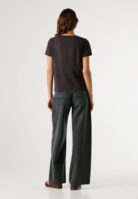 Γυναίκα στέκεται με πλάτη, φορώντας ένα μαύρο κοντομάνικο t-shirt, ψηλόμεσα φαρδιά τζιν παντελόνια και σκούρα κόκκινες αθλητικές έλσες.