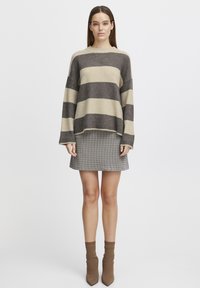 Pull en tricot surdimensionné avec des rayures marron foncé et beige, associé à une jupe à carreaux grise et des chaussettes cheville beige clair, chaussures plates non visibles.