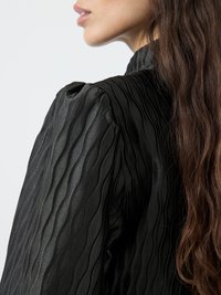 Black textured blouse with a wavy design in satin fabric. Features puffed shoulders and a high neckline. Smooth finish and sleek silhouette.