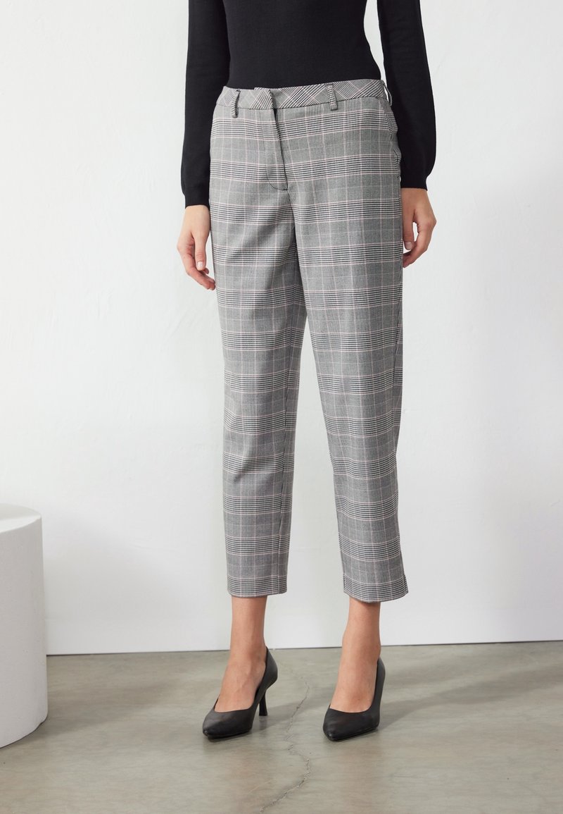 Femme portant un pantalon droit gris à carreaux et des talons noirs, les mains détendues le long du corps, debout sur un sol en béton poli.