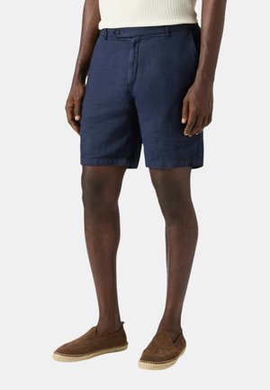 Uomo con pantaloncini blu navy, top bianco senza maniche e scarpe marroni slip-on, in piedi davanti a uno sfondo chiaro e uniforme.