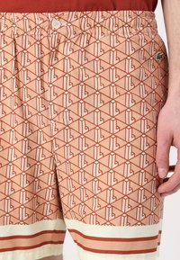 Menns badeshorts i lyse korall med et geometrisk mønster i rødt. Har elastisk liv, knappelukking og kremfargede striper nederst.