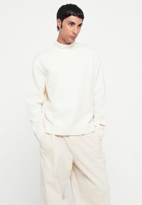 Suéter blanco de punto con un cuello alto y acanalado. Textura suave y corte relajado, combinado con pantalones sueltos de color beige claro. Detalle sutil de marca.