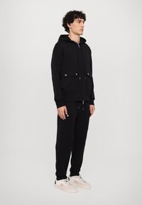 Sudadera negra con cremallera y bolsillos frontales, combinada con pantalones de chándal a juego y zapatillas de color claro. Textura de tela suave.