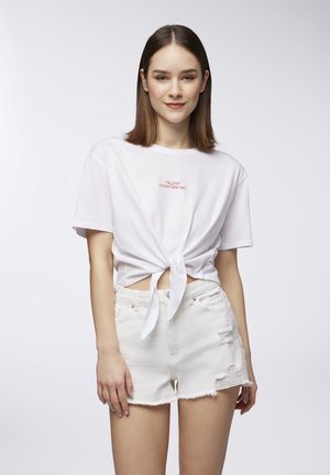 T-shirt bianco a maniche corte con nodo davanti, con testo rosso "TALENTO SOPRA IL RIPENSARE," abbinato a pantaloni corti bianchi strappati.