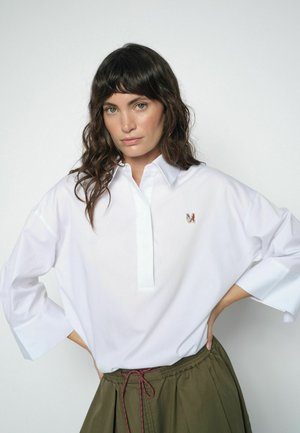 Kobieta o falowanych ciemnych włosach, ubrana w białą koszulę z kołnierzykiem z małym kolorowym logo "M" oraz oliwkowo-zielone spodnie z czerwonym sznurkiem.