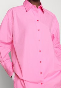 Rosa Hemd mit Knopfleiste, spitzem Kragen, langen Ärmeln und sichtbaren roten Knöpfen vorne. Hergestellt aus einem glatten, leichten Stoff.