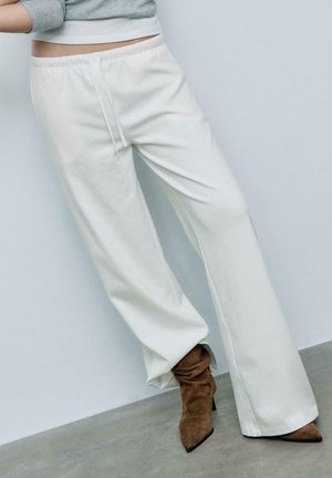 Osoba nosiaca biele nohavice na sťahovanie a hnedé semišové topánky na podpätku so špicatou špičkou na pozadí obyčajnej steny.