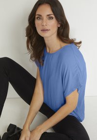 Top blu corto a maniche corte realizzato in un tessuto testurizzato, con scollatura rotonda, abbinato a pantaloni neri attillati. Il modello è seduto su una superficie chiara.