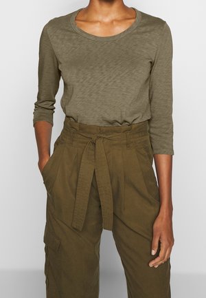 Personne portant une chemise à manches longues vert olive rentrée dans un pantalon taille haute vert olive avec une ceinture en tissu nouée à la taille.