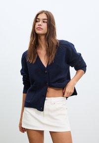 Kornblumenblauer, kurz geschnittener Cardigan aus strukturiertem Strick, mit tiefem V-Ausschnitt, vorderem Knopfverschluss und zwei vorderen Taschen. Kombiniert mit einem weißen Minirock.