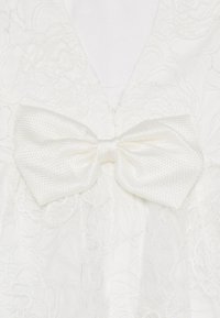 Weißes Kleid mit floraler Spitze und einer auffälligen, strukturierten weißen Satin-Schleife in der Mitte, die einen eleganten, geschichteten Look kreiert.