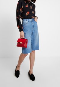 Donna che indossa una blusa nera floreale, shorts in denim fino al ginocchio, mocassini neri, e tiene in mano una piccola borsa rossa su sfondo bianco.