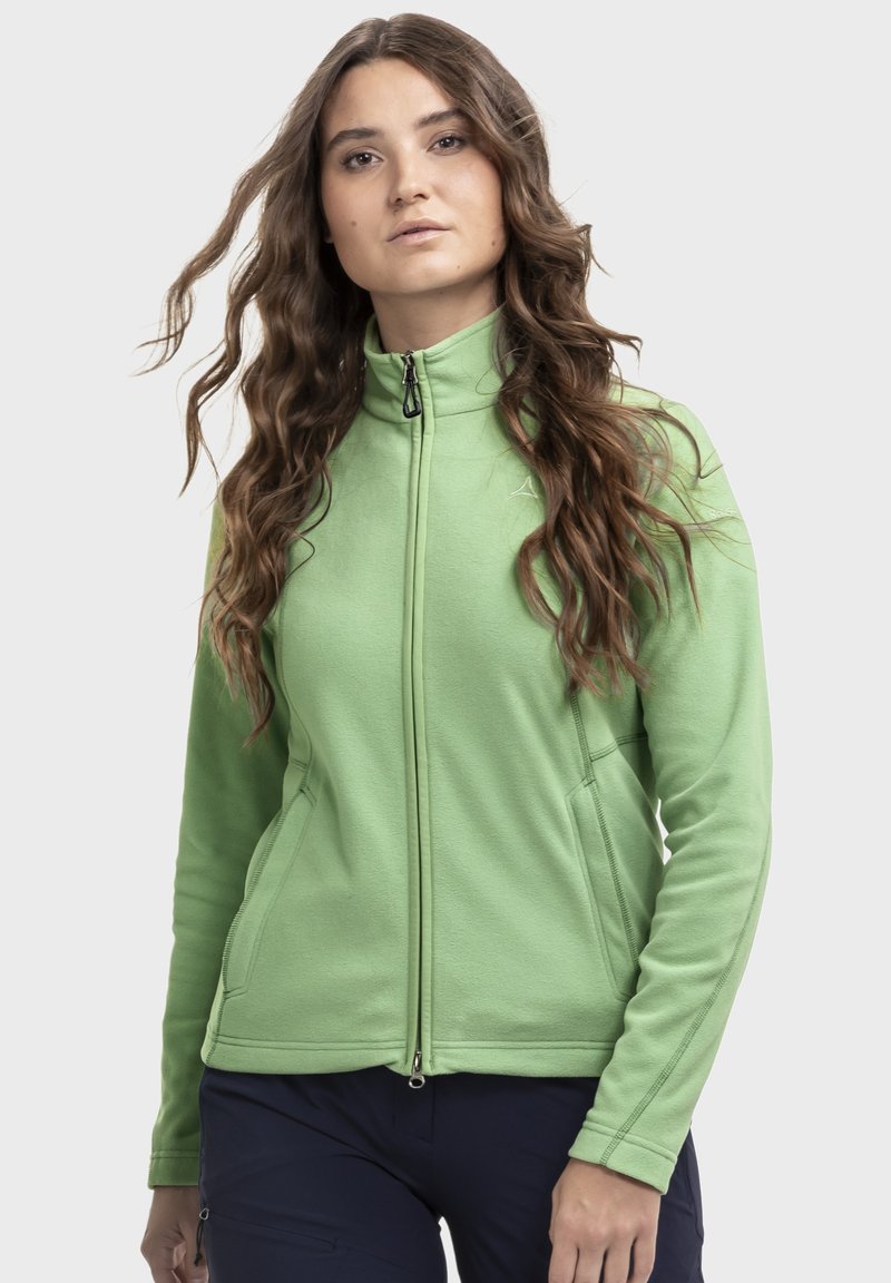 Frau mit langen, welligen braunen Haaren, die eine hellgrüne Reißverschlussjacke und dunkle Hosen trägt, vor einem einfarbigen hellen Hintergrund.