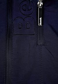 Tmavě modrá bunda na zip s texturovanou látkou, zdobená vyraženým logem "Be" a značkovým jezdcem zipu s nápisem "Bench." výrazně zobrazeným.