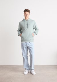 Felpa verde chiaro con tasca frontale e coulisse, abbinata a jeans azzurri e sneakers bianche. Design semplice senza motivi o dettagli.