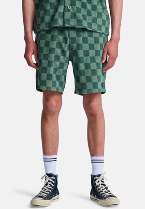 Man draagt groene geruite shorts, bijpassend geruit overhemd, witte kousen met zwarte strepen en zwarte hoge sneakers met witte veters.