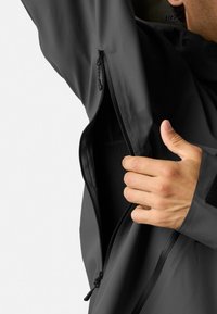 Grå jakke med en lynlås lomme og velcro-manchet. Materialet ser glat ud. Hånd vist holder i lynlåsen, hvilket afslører en mørk inderside.