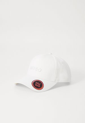 Witte baseballcap met geborduurd "HUGO" logo in verhoogde letters. Heeft een ronde vorm en een ronde hangtag met zwarte en rode accenten.