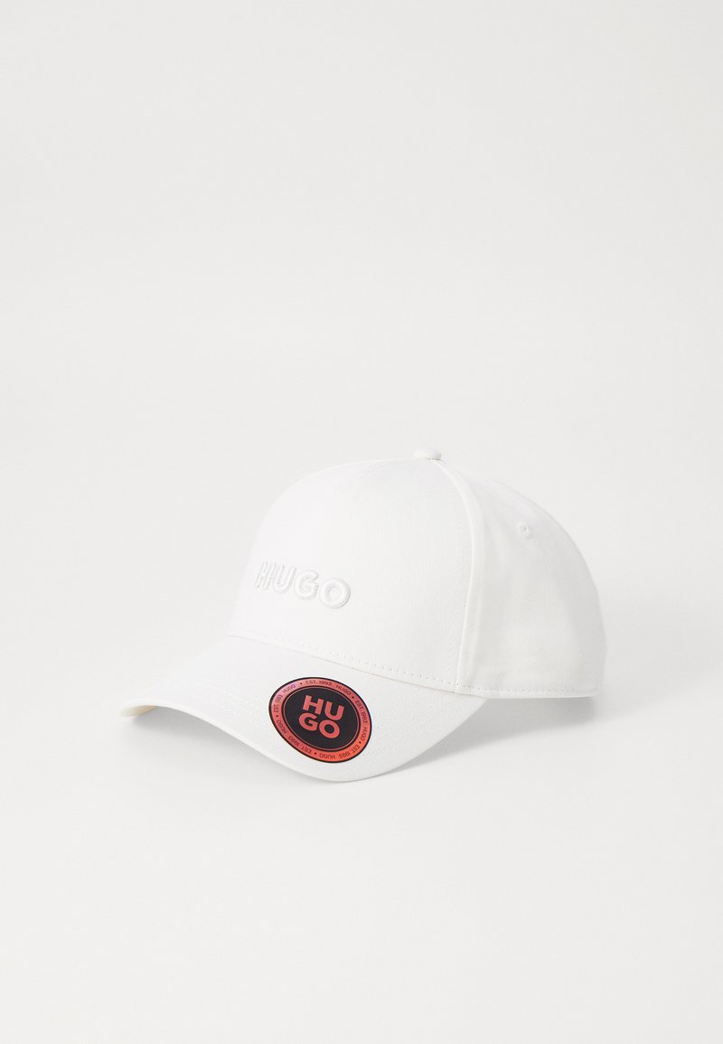 Hvid baseball cap med broderet "HUGO" logo i forhøjede bogstaver. Har en rund form og et rundt mærke med sorte og røde accenter.