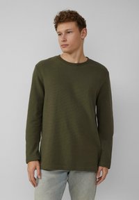 Olijfgroene langemouwen shirt van een gestructureerde stof, met een ronde hals en rechte zoom, gedragen met lichtblauwe jeans.
