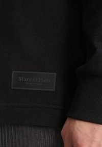 Schwarzer Sweatshirt mit gerippten Bündchen, versehen mit einem schwarzen Stoffetikett mit eingeprägtem Text "Marc O'Polo EST. IN STOCKHOLM." Die Textur wirkt glatt.