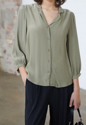 Woman wearing a light olive green button-up blouse with puffed sleeves and black pleated pants, holding a small black handbag.