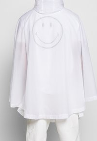 Vit huva poncho med avslappnad passform, som har en stor, grå smiley-design på ryggen. Tillverkad av lätt och mjukt material.