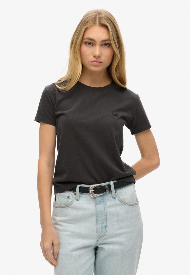 T-shirt nera in cotone con maniche corte, scollatura rotonda, con un piccolo logo sul lato sinistro del petto. Indossata con jeans azzurri chiari e una cintura nera.