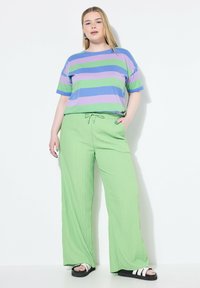 Gestreept kortmouwen top in paars, groen en blauw; losse groene broek met trekkoord en structuur; zwarte sandalen met witte strepen.