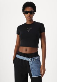 Črna skrajšana majica s kratkimi rokavi z zlatim logotipom "GUESS", v kombinaciji s črnimi trenirkami, ki vključujejo denim panel v pasu in elastičnega vrvico za prilagajanje.