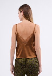 Top de satén marrón con tirantes finos y un escote en V decorado con encaje. Combinado con pantalones verdes estampados con manchas negras y naranjas.