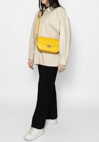 Camicia beige oversize, pantaloni neri a gamba larga e sneakers bianchi. Una borsa a tracolla gialla presenta una texture liscia e un dettaglio con chiusura metallica.