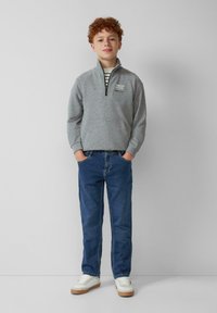 Sudadera gris con cremallera en el cuello, con un bolsillo frontal. Jeans de mezclilla azules y zapatillas blancas con acentos beige completan el look.