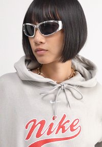 Pelēka kapuce ar sarkanu Nike logo, ar aukliņu ap kaklu. Cilvēks valkā baltas kaķa acu saulesbrilles un zelta ķēdes kaklarotu.