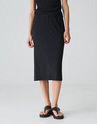 Schwarzer, strukturierter Midirock mit einem gerafften Bund. Kombiniert mit flachen, offenen Sandalen mit einem Schnallendetail. Einfaches, elegantes Design.