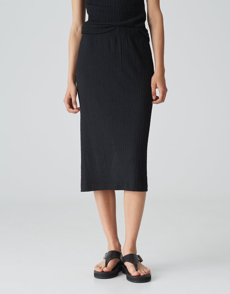 Schwarzer, strukturierter Midirock mit einem gerafften Bund. Kombiniert mit flachen, offenen Sandalen mit einem Schnallendetail. Einfaches, elegantes Design.