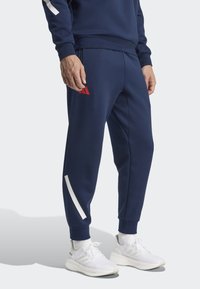 Navy blue athletic joggers with white accents and red logo. Features elastic waistband and tapered legs, paired with white athletic shoes.