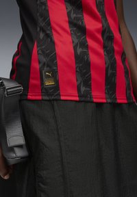 Maillot de football noir et rouge à rayures, fabriqué en tissu texturé, avec un logo doré et des détails imprimés, associé à une bandoulière noire.