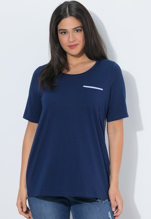 Kvinde med langt mørkt hår iført en marineblå kortærmet skjorte med brystlomme og slidte blå jeans, stående mod en hvid baggrund.