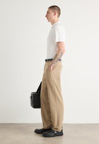 Beige wijde pijpenbroek, witte poloshirt met korte mouwen, zwarte riem en zwarte schoenen. Houdt een zwarte tas vast. Heeft een tatoeage op de linkerarm.