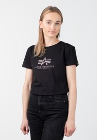 Juoda medvilninė marškinėliai su metaliniu logotipu ant krūtinės, trumpomis rankovėmis ir klasikine apvalia kaklo išpjova. Sukombinuoti su tamsiomis džinsais.