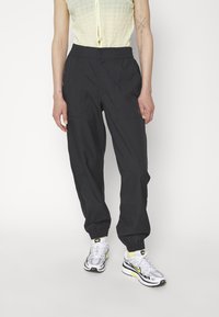 Persona que lleva pantalones elásticos negros con cintura y tobillo, zapatillas Nike blancas y amarillas, y una blusa sin mangas beige texturizada, de pie contra un fondo blanco.