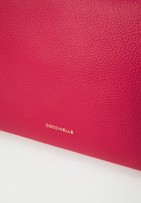 Rdeča usnjena torbica z teksturirano površino. Zlati logotip "COCCINELLE" je izpostavljen na spodnjem desnem delu.