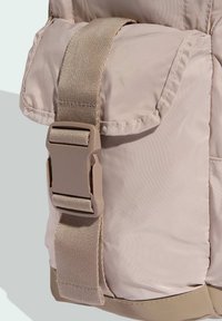 Svetlo béžová taška z nylonu s textúrovaným povrchom, vybavená veľkou prednou kapsou a nastaviteľným béžovým popruhom s plastovou prackou.