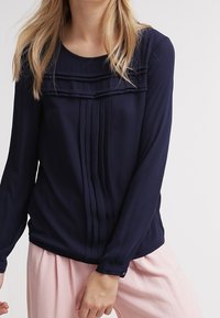 Blusa a maniche lunghe blu navy con pieghe e dettagli a strati lungo il colletto, realizzata in un tessuto leggero e morbido.